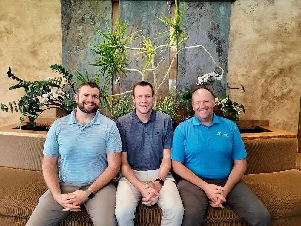 Three adults sitting on a bench in front of indoor plants, one wearing an Oak Mountain Dental shirt.