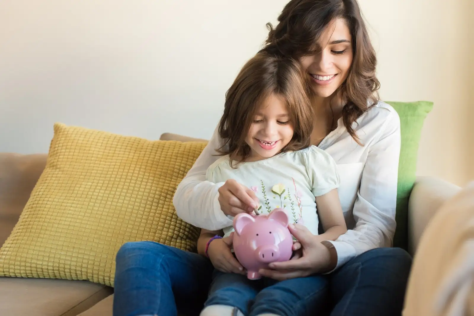 An adult and a child sitting on a couch putting a coin into a pink piggy bank.